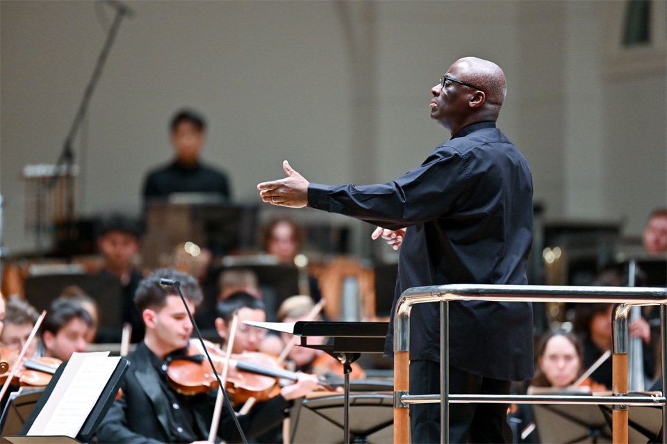 A black man wearing formal attire, conducting an orchestra, with students performing on their instruments, also wearing formal attire